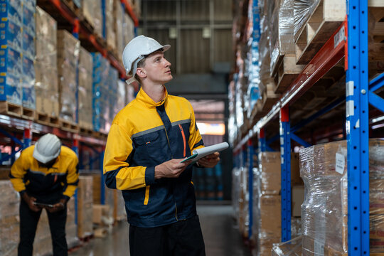 Man warehouse worker wearing uniform and helmet safety standing with clipboard for checking stock products on shelves in warehouse. Logistics, Distribution Center concept