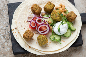Wheat wraps with falafel, fresh vegetables and yogurt on a black wooden serving board, horizontal shot, middle closeup, selective focus