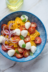 Salad with corn, red cherry tomatoes and mini mozzarella cheese served in a blue bowl, vertical shot on a white stone background, middle close-up