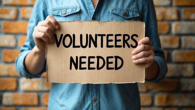 Man holding a cardboard sign with the words "Volunteers Needed"