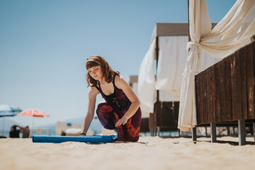 A woman is placing a blue yoga mat on sandy beach near shaded cabanas preparing for relaxation or exercise under a sunny sky along the coastline.