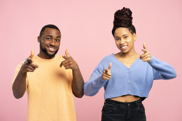 Cheerful African American man and woman pointing fingers at camera on pink background