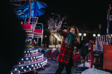 A cheerful woman wearing a scarf and coat throws snow joyfully in a festive outdoor setting. The background shows a colorful park illuminated by lights, suggesting a lively celebration and winter