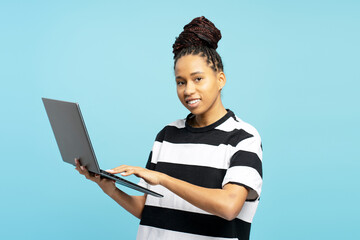 Attractive African American woman typing on laptop isolated blue background, looking at camera