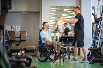 Encouraging personal trainer give high five, supports glad wheelchair athlete on rehabilitation...