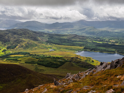 A mountaintop scene overlooking a valley. The valley contains a lake and a mix of green fields, forests, and rocky outcrops. The sky is cloudy.