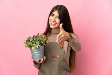 Young gardener girl holding a plant isolated on pink background shaking hands for closing a good...