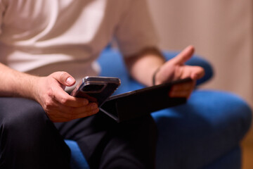 A person is seen sitting comfortably on a blue couch while using both a smartphone and a tablet