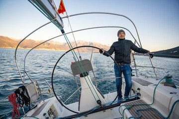 Young man captain standing at the helm and controls a sailboat during a journey by sea in winter...