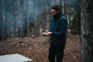 A man holds a piece of wood while preparing a campfire in a dense forest, portraying a rustic...