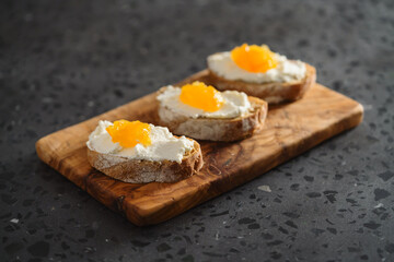 Three ciabatta slices with ricotta and orange marmalade on olive wood board on terrazzo surface