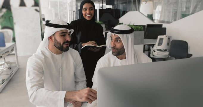 Three Arabian teammates dressed in traditional kandura and hijab attires discuss task using modern computer, collaborate on online project, review update or presentation gathered together in coworking