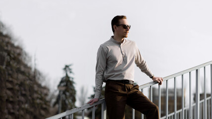 Attractive redhead Caucasian young man in stylish clothes poses while walking outdoors on a spring or autumn day. Confident man wearing sunglasses. Lifestyle, career, travel.