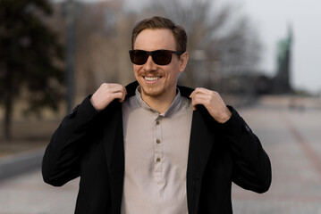 Obraz premium Portrait of attractive redhead Caucasian young man wearing sunglasses and black jacket. Happy and confident man walking in a park on an autumn or spring day. Lifestyle, positivity.