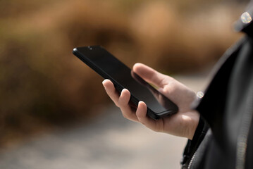 Close-up of hands of a young woman in a leather jacket using a mobile phone while walking outdoors on a spring or autumn day. Technology, network, business, social media.