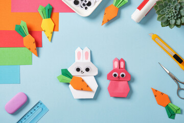 Top view of the creative desktop. Pink and white origami bunnies with orange carrots. Diy art supplies: paper, scissors, stationery knife, markers