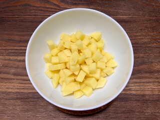 Fresh peeled potatoes that have been cut into small pieces, placed on a white ceramic bowl on a wooden table