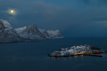 Obraz premium Husøy auf der Insel Senja in Norwegen zur blauen Stunde bei klarer Sicht mit Blick auf leuchtende Häuser im verschneiten Fjord und einem hellen Vollmond über den Bergen am ruhigen Morgenhimmel
