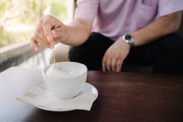 A hand holding a spoon with creamy milk foam above a white cup on a saucer with a napkin, capturing the elegance and warmth of a relaxing coffee break. Perfect for beverage and lifestyle themes.