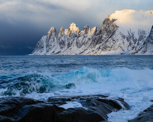Die Okshornan auf Senja in Norwegen zur goldenen Stunde, im Licht leuchtend vor einem aufziehenden dunklen Sturmhimmel, während hohe Wellen im Vordergrund das aufgewühlte Meer bestimmen