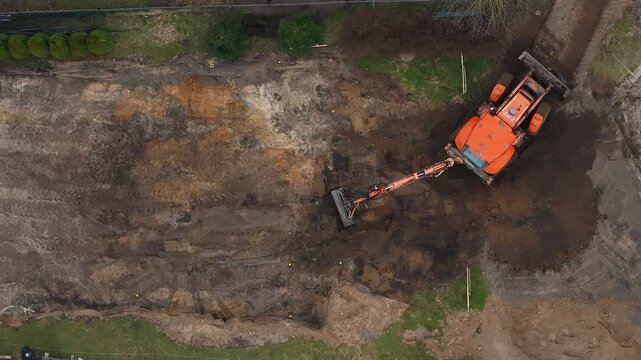 An aerial view shows an orange excavator working on a construction site, bordered by grass and sparse vegetation, with visible fencing and materials.