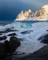 Die Okshornan auf Senja in Norwegen zur goldenen Stunde, im Licht leuchtend vor einem aufziehenden dunklen Sturmhimmel, während hohe Wellen im Vordergrund das aufgewühlte Meer bestimmen