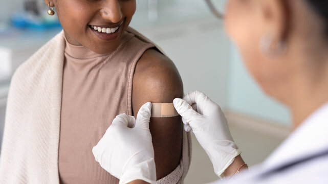 Female doctor wearing protective rubber gloves sticking adhesive plaster on shoulder of happy Black patient woman visiting clinic for vaccination, protecting place of shot from infection. Banner image
