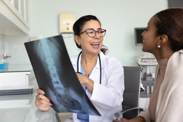 Cheerful senior Caucasian doctor and young African patient woman discussing roentgen image of spine and pelvic bones, talking on consultation meeting, smiling, laughing