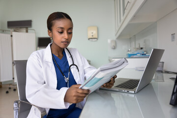 Serious young African doctor woman reading large stack of paper medical records at workplace table,...