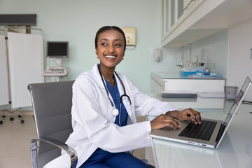 Cheerful young African American medical therapist working at laptop in hospital examination room,...