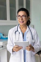 Positive middle aged doctor woman in glasses holding tablet pad, standing in clinic work office, using modern technology, health care application on digital device. Vertical shot