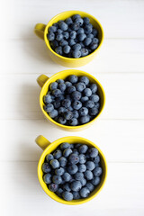 Fresh juicy wild blueberries in three yellow mugs close-up on white table surface, top view, soft focus