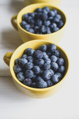 Fresh juicy wild blueberries in yellow mugs close-up on white table surface, top view, soft focus