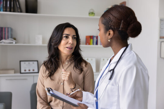 Mature Caucasian woman visiting young African female doctor for consultation, telling about healthcare, sickness symptoms. Physician filling medical form, listening to patient in clinic office
