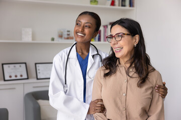 Happy young Black doctor and senior woman looking away, laughing, discussing successful treatment,...