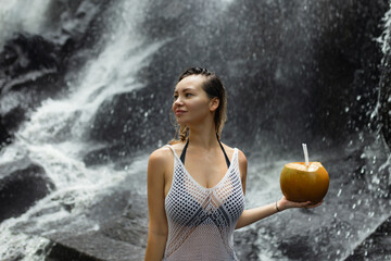 Beautiful sexy woman stands near a waterfall in Bali and holds a coconut in her hand
