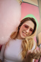 Naklejka premium Young woman wearing sunglasses smiles while holding cotton candy near a colorful mural. The bright colors and joyful expression capture a lively moment outdoors.
