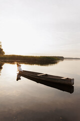 A woman in a white dress sits in a boat on a lake at sunset, dipping her hand into the water. Warm...