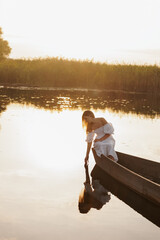 A woman in a white dress sits in a boat on a lake at sunset, dipping her hand into the water. Warm...