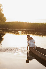 A woman in a white dress sits in a boat on a lake at sunset, dipping her hand into the water. Warm...