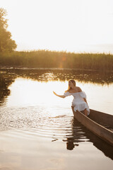 A woman in a white dress sits in a boat on a lake at sunset, dipping her hand into the water. Warm...