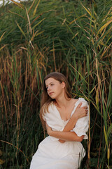 A young woman in a white dress sits barefoot in a boat among reeds on a lake. She gazes to the side, creating a serene and nature-connected atmosphere. Warm evening light enhances the harmony