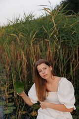 A young woman in a white dress sits barefoot in a boat among reeds on a lake. She gazes to the side, creating a serene and nature-connected atmosphere. Warm evening light enhances the harmony