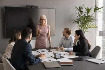 Mature Caucasian woman business coach makes speech stand in front of employees, share experience, skills during corporate training in office. Communication between business partners or shareholders