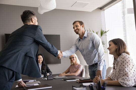 Two smiling male colleagues shake hands, closing profitable commercial deal at meeting with employees in office. Businessmen handshaking get acquainted, greeting partner, making agreement. Partnership