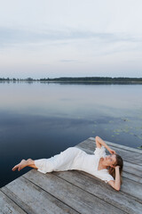 A woman in a white dress lies on a wooden pier near a lake covered with water lilies. The atmosphere is peaceful in harmony with nature. The dark water reflects the sky. The light romantic dress 