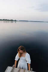 Fototapeta premium A woman in a white dress stands in the water near a wooden pier, holding onto the edge. The evening sky reflects in the calm lake. In the distance, a boat with people adds to the sense of solitude