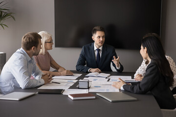 Serious young professional businessman speaking at team meeting, presenting creative idea, take part in brainstorming session, telling project plan or strategy, talking to diverse teammates in office