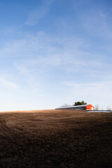 A New England Farm with a red barn sitting on the top of a hill with a hay field in the foreground. The sky is blue with whispy clouds and golden hour is creating nice light on the field and barn.
