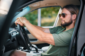 Naklejka premium Young man with sunglasses sitting in a car, focused on driving, showcasing a calm demeanor against a blurred green background